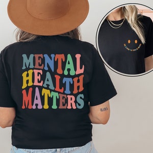 May include: Black t-shirt with the text "MENTAL HEALTH MATTERS" in rainbow colors. The text is in a retro font style. The back of the shirt is shown. A second black t-shirt is shown in a mirror reflection with the text "Be kind to your mind" and a smiley face.