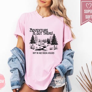 May include: A light pink t-shirt with the text "Adventure is out there! But so are serial killers" in black. The graphic features a winding path through a forest. The shirt is labeled "Super Soft".