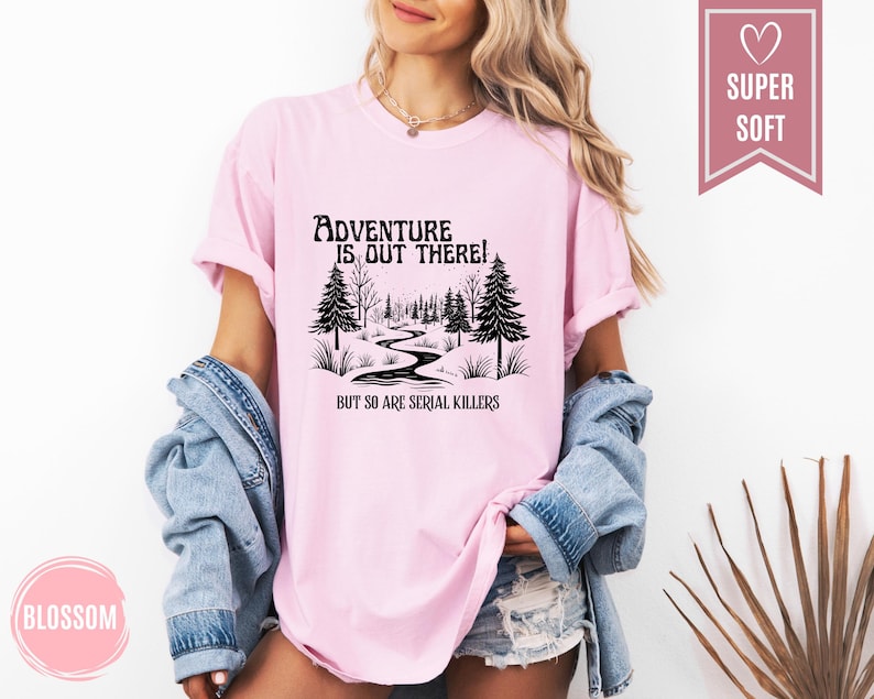 May include: A light pink t-shirt with the text "Adventure is out there! But so are serial killers" in black. The graphic features a winding path through a forest. The shirt is labeled "Super Soft".