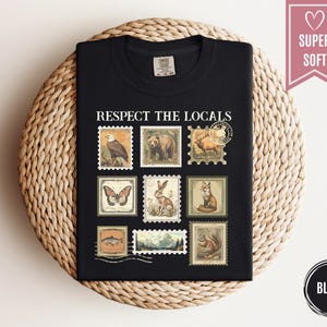 May include: Black t-shirt with the phrase "RESPECT THE LOCALS" and vintage-style postage stamp graphics of various animals. The stamps include an eagle, bear, deer, butterfly, rabbit, fox, fish, mountain scene, and squirrel.