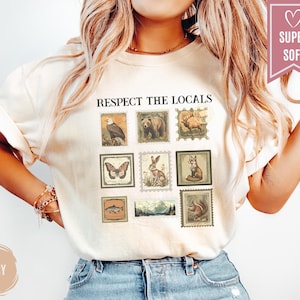 May include: Ivory-colored t-shirt with the text "RESPECT THE LOCALS" above a series of vintage-style postage stamp illustrations featuring an eagle, bear, deer, butterfly, rabbit, fox, fish, landscape, and squirrel. The shirt has a relaxed fit.