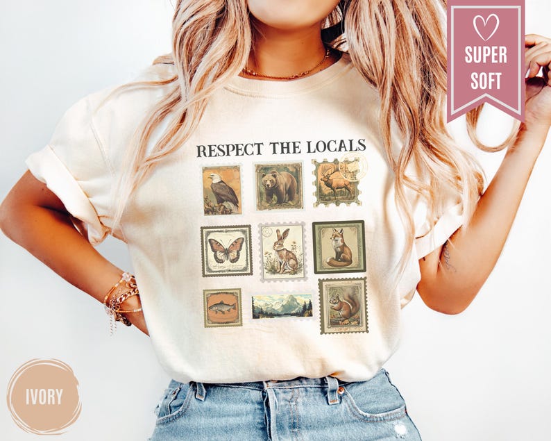 May include: Ivory-colored t-shirt with the text "RESPECT THE LOCALS" above a series of vintage-style postage stamp illustrations featuring an eagle, bear, deer, butterfly, rabbit, fox, fish, landscape, and squirrel. The shirt has a relaxed fit.