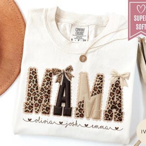 May include: Ivory-colored t-shirt with the word "MAMA" in a mix of leopard print and beige fabric. The letters are embellished with bows. Below the word are the names "olivia", "josh", and "emma". A gold necklace is visible.