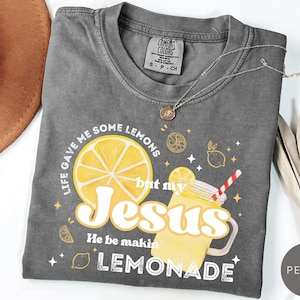 Puede incluir: Una camiseta gris Comfort Colors con el texto "Life gave me some lemons but my Jesus He be makin' lemonade." El gr&aacute;fico incluye una gran rodaja de lim&oacute;n amarillo y una bebida de limonada. Un collar plateado est&aacute; sobre la camiseta.