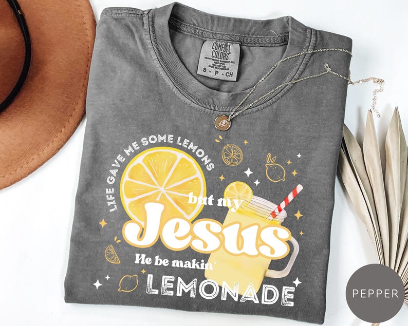 Puede incluir: Una camiseta gris Comfort Colors con el texto "Life gave me some lemons but my Jesus He be makin' lemonade." El gr&aacute;fico incluye una gran rodaja de lim&oacute;n amarillo y una bebida de limonada. Un collar plateado est&aacute; sobre la camiseta.