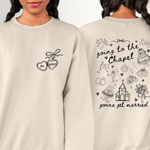 K&ouml;nnte beinhalten: Cremefarbenes Sweatshirt mit schwarzer Strichzeichnung. Die Vorderseite zeigt zwei herzf&ouml;rmige Anh&auml;nger mit dem Text "Mrs. Wilson". Die R&uuml;ckseite zeigt Illustrationen zum Thema Hochzeit und die S&auml;tze "going to the Chapel" und "gonna get married".