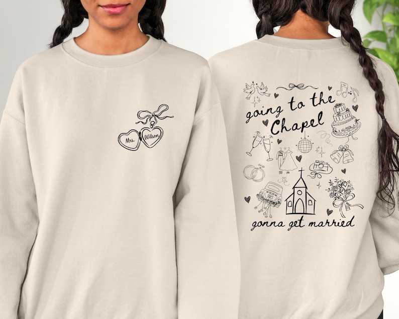 K&ouml;nnte beinhalten: Cremefarbenes Sweatshirt mit schwarzer Strichzeichnung. Die Vorderseite zeigt zwei herzf&ouml;rmige Anh&auml;nger mit dem Text "Mrs. Wilson". Die R&uuml;ckseite zeigt Illustrationen zum Thema Hochzeit und die S&auml;tze "going to the Chapel" und "gonna get married".
