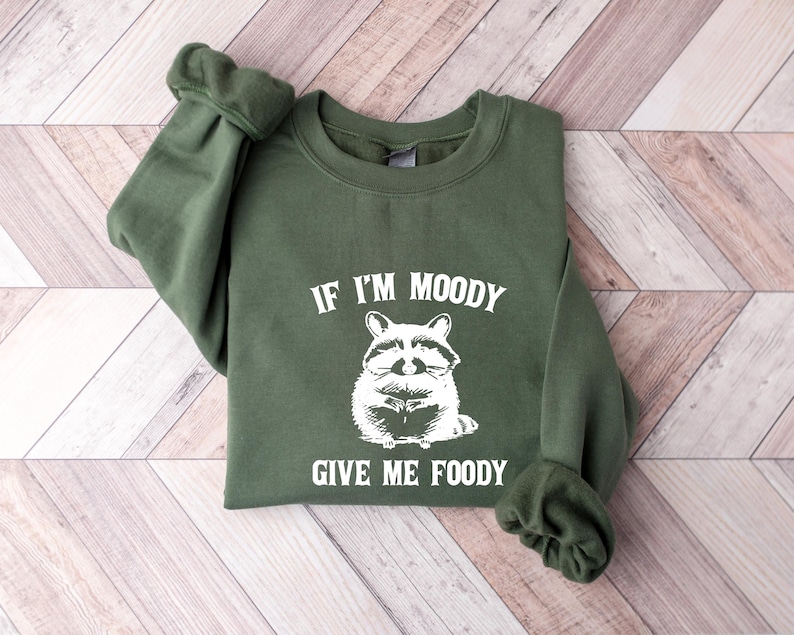 May include: Green sweatshirt with a white graphic of a raccoon and the text "If I'm Moody Give Me Foody".