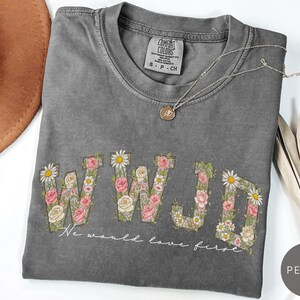 K&ouml;nnte beinhalten: Pfefferfarbenes T-Shirt mit floralem "WWJD"-Design. Die Buchstaben sind mit rosa Rosen, wei&szlig;en G&auml;nsebl&uuml;mchen und Gr&uuml;n gef&uuml;llt. Der Text "We would love first" steht darunter. Eine silberne Halskette mit Anh&auml;nger befindet sich auf dem Shirt.