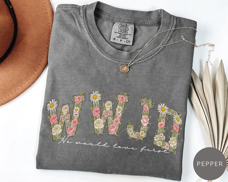 K&ouml;nnte beinhalten: Pfefferfarbenes T-Shirt mit floralem "WWJD"-Design. Die Buchstaben sind mit rosa Rosen, wei&szlig;en G&auml;nsebl&uuml;mchen und Gr&uuml;n gef&uuml;llt. Der Text "We would love first" steht darunter. Eine silberne Halskette mit Anh&auml;nger befindet sich auf dem Shirt.