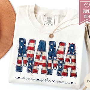 May include: Ivory-colored t-shirt with the word "MAMA" in a patriotic design, featuring red and white stripes and blue stars. The names "olivia", "josh", and "emma" are printed below. A gold necklace and a "Super Soft" label are also visible.