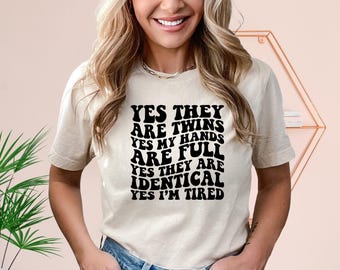 May include: A beige t-shirt with black text that reads "YES THEY ARE TWINS YES MY HANDS ARE FULL YES THEY ARE IDENTICAL YES I'M TIRED"
