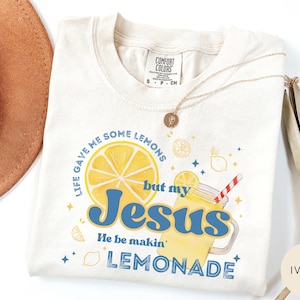 Puede incluir: Camiseta de color marfil con la frase "Life gave me some lemons but my Jesus He be makin' lemonade" en azul y amarillo. El dise&ntilde;o incluye una rodaja de lim&oacute;n, un vaso de limonada y estrellas. La camiseta es de la marca Comfort Colors.