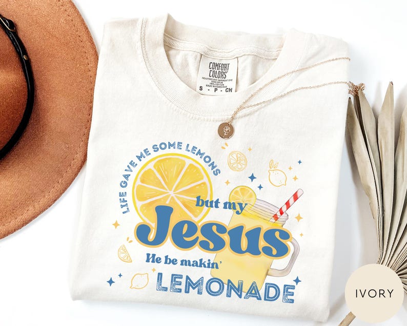 Puede incluir: Camiseta de color marfil con la frase "Life gave me some lemons but my Jesus He be makin' lemonade" en azul y amarillo. El dise&ntilde;o incluye una rodaja de lim&oacute;n, un vaso de limonada y estrellas. La camiseta es de la marca Comfort Colors.