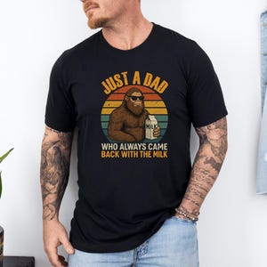May include: Black t-shirt with a graphic of a Bigfoot wearing sunglasses, holding a milk carton, and the text "JUST A DAD WHO ALWAYS CAME BACK WITH THE MILK" in orange and white. The design is set against a retro sunset.