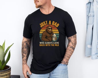 May include: Black t-shirt with a graphic of a Bigfoot wearing sunglasses, holding a milk carton, and the text "JUST A DAD WHO ALWAYS CAME BACK WITH THE MILK" in orange and white. The design is set against a retro sunset.