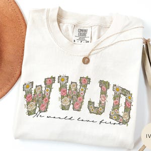 K&ouml;nnte beinhalten: Elfenbeinfarbenes Comfort Colors T-Shirt mit den Buchstaben "WWJD" in einem floralen Design aus rosa Rosen, wei&szlig;en G&auml;nsebl&uuml;mchen und gr&uuml;nen Bl&auml;ttern. Der Satz "We would love first" steht darunter. Eine goldene Halskette liegt auf dem Shirt.