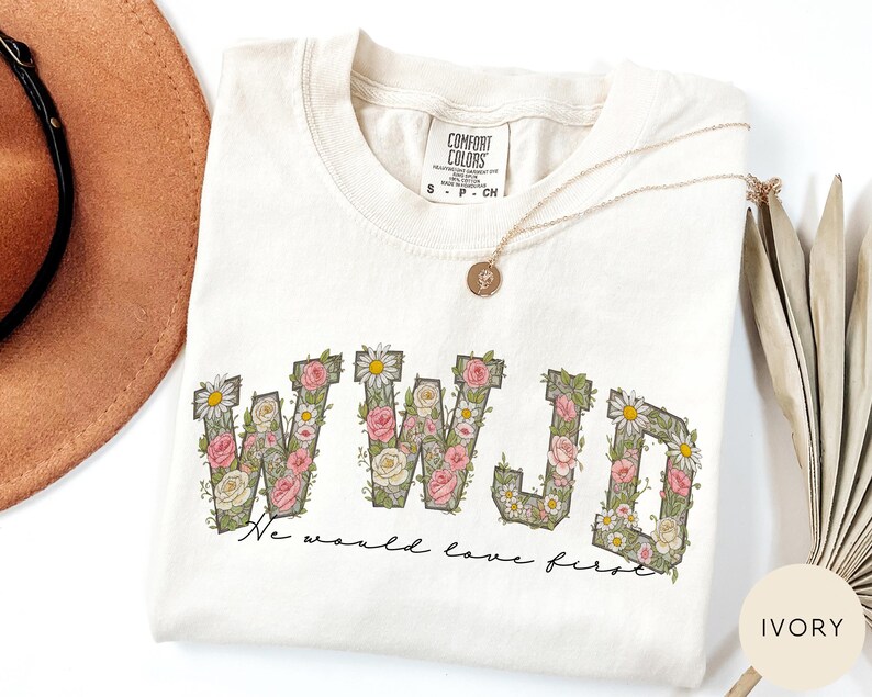 K&ouml;nnte beinhalten: Elfenbeinfarbenes Comfort Colors T-Shirt mit den Buchstaben "WWJD" in einem floralen Design aus rosa Rosen, wei&szlig;en G&auml;nsebl&uuml;mchen und gr&uuml;nen Bl&auml;ttern. Der Satz "We would love first" steht darunter. Eine goldene Halskette liegt auf dem Shirt.