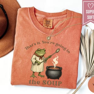 May include: A coral-colored t-shirt featuring a cartoon frog character stirring a pot of soup. The text on the shirt reads "That's it. You're going in the soup." A gold necklace and a brown hat are also visible.
