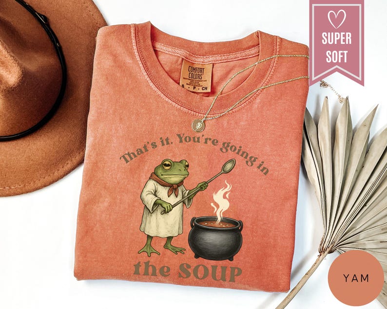 May include: A coral-colored t-shirt featuring a cartoon frog character stirring a pot of soup. The text on the shirt reads "That's it. You're going in the soup." A gold necklace and a brown hat are also visible.