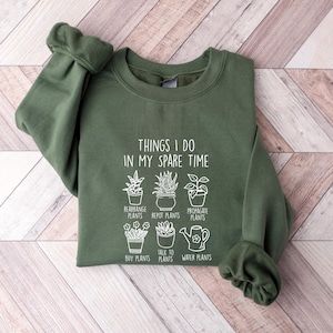 May include: A green sweatshirt with a white graphic that says "Things I do in my spare time" and shows a series of potted plants with the words "Rearrange Plants", "Repot Plants", "Propagate Plants", "Buy Plants", "Talk to Plants", and "Water Plants".