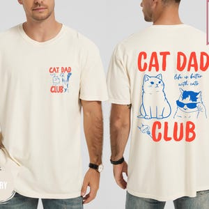 May include: Ivory-colored t-shirt with "CAT DAD CLUB" text and cat illustrations. The back features a larger design with the text "Life is better with cats." The shirt is labeled "SUPER SOFT."