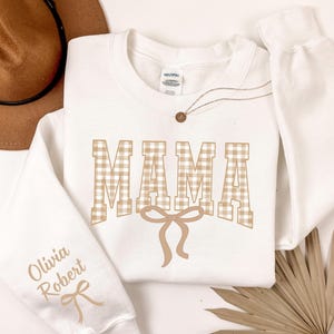 May include: A white sweatshirt with the word "MAMA" in a tan gingham pattern. A tan bow is below the word. The sleeve has the names "Olivia Robert" printed in tan. A brown hat and necklace are also in the image.
