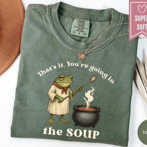 May include: Moss green t-shirt with a frog chef illustration holding a spoon over a pot of soup. The text "That's it. You're going in the SOUP" is printed on the shirt. The shirt has a crew neck and short sleeves.