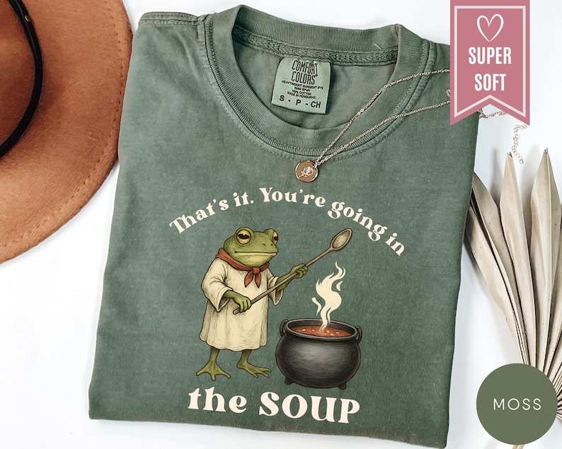 May include: Moss green t-shirt with a frog chef illustration holding a spoon over a pot of soup. The text "That's it. You're going in the SOUP" is printed on the shirt. The shirt has a crew neck and short sleeves.