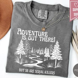 May include: A gray t-shirt with the text "Adventure is Out There! But So Are Serial Killers" in white. The graphic features a forest scene with trees and a winding river. A silver necklace with a pendant rests on the shirt.