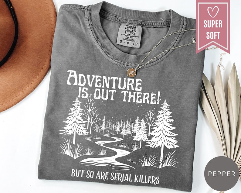 May include: A gray t-shirt with the text "Adventure is Out There! But So Are Serial Killers" in white. The graphic features a forest scene with trees and a winding river. A silver necklace with a pendant rests on the shirt.