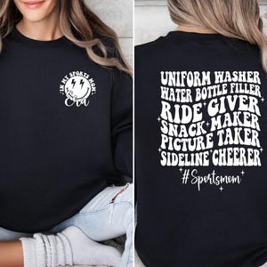 Pode incluir: Camisola de moletom preta com texto branco que diz "Uniform Washer, Water Bottle Filler, Ride Giver, Snack Maker, Picture Taker, Sideline Cheerer #Sportsmom" nas costas e um rosto sorridente com raios e o texto "In My Sports Mom Era" na frente.