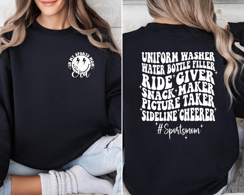 Pode incluir: Camisola de moletom preta com texto branco que diz "Uniform Washer, Water Bottle Filler, Ride Giver, Snack Maker, Picture Taker, Sideline Cheerer #Sportsmom" nas costas e um rosto sorridente com raios e o texto "In My Sports Mom Era" na frente.