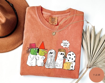 May include: Coral-colored Comfort Colors t-shirt featuring a cartoon of Halloween characters, including a witch, a wizard, and ghosts. One ghost holds a box, and a thought bubble says, "I GOT A ROCK." A gold necklace and a brown hat are also visible.