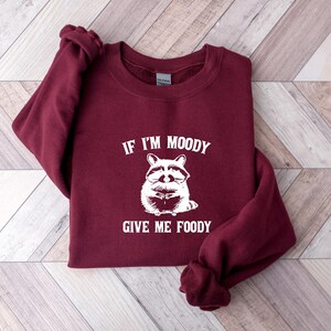May include: Burgundy sweatshirt with a white raccoon graphic and the text "If I'm Moody Give Me Foody".