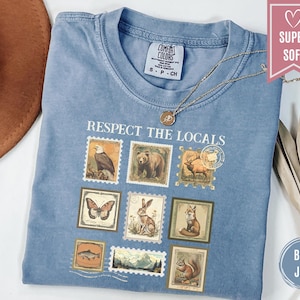 May include: Blue jean colored t-shirt with the phrase "Respect the Locals" printed on the front. The design features postage stamp-style illustrations of various animals, including a bear, eagle, and fox. A gold necklace rests on the shirt.