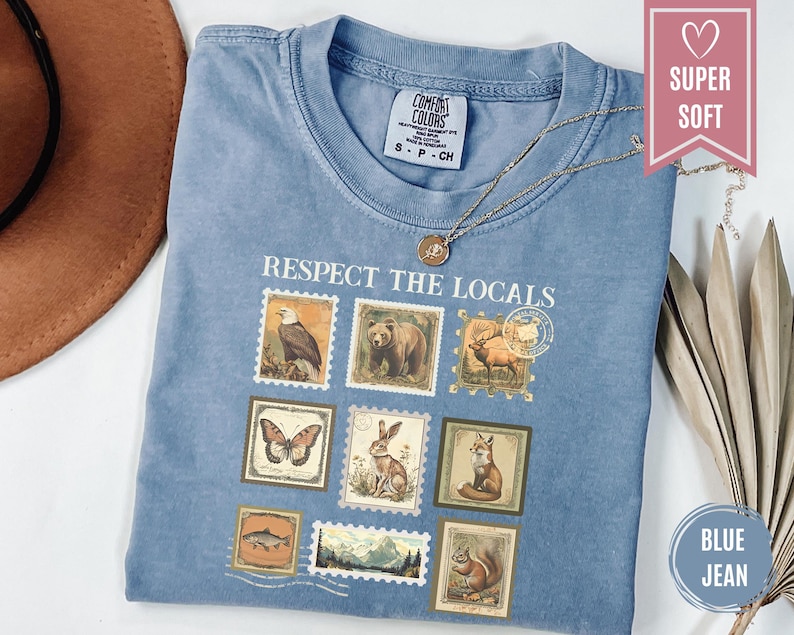 May include: Blue jean colored t-shirt with the phrase "Respect the Locals" printed on the front. The design features postage stamp-style illustrations of various animals, including a bear, eagle, and fox. A gold necklace rests on the shirt.