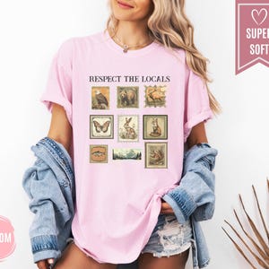 May include: A light pink t-shirt with the text "RESPECT THE LOCALS" and a collection of vintage-style postage stamp illustrations featuring various animals and landscapes. The shirt is made of a soft material.