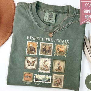 May include: A moss green t-shirt with the text "Respect the Locals" and postage stamp-style illustrations of wildlife. The shirt features an eagle, bear, deer, butterfly, rabbit, fox, fish, squirrel, and mountain scene. A gold necklace is draped on the shirt.
