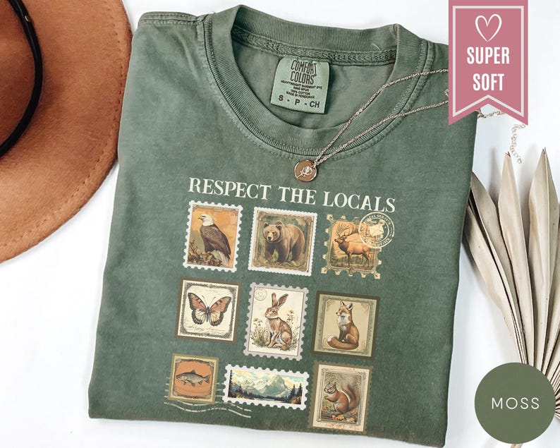 May include: A moss green t-shirt with the text "Respect the Locals" and postage stamp-style illustrations of wildlife. The shirt features an eagle, bear, deer, butterfly, rabbit, fox, fish, squirrel, and mountain scene. A gold necklace is draped on the shirt.