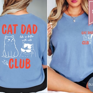 May include: Blue jean colored t-shirts with the text "CAT DAD CLUB" in red. The back of the shirt features a cat illustration and the phrase "life is better with cats." The front of the shirt has a smaller "CAT DAD CLUB" design.