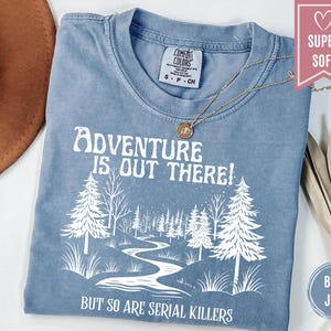 May include: Blue jean colored t-shirt with the text "Adventure is out there! But so are serial killers" in white. The shirt features a graphic of a river winding through a forest. A gold necklace rests on the shirt.