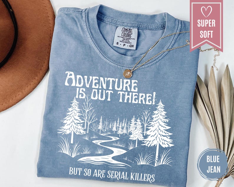 May include: Blue jean colored t-shirt with the text "Adventure is out there! But so are serial killers" in white. The shirt features a graphic of a river winding through a forest. A gold necklace rests on the shirt.