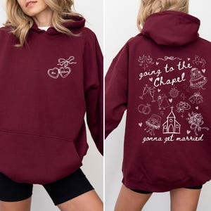 K&ouml;nnte beinhalten: Ein weinroter Kapuzenpullover mit wei&szlig;en grafischen Texten und Illustrationen. Die Vorderseite zeigt zwei herzf&ouml;rmige Designs mit dem Text "Mrs. [Name]" im Inneren. Auf der R&uuml;ckseite steht der Satz "going to the Chapel gonna get married" mit Illustrationen zum Thema Hochzeit.
