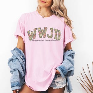 K&ouml;nnte beinhalten: Hellrosa T-Shirt mit den Buchstaben "WWJD" in floralem Design. Darunter steht der Text "No world love first". Das Shirt wird mit einer Jeansjacke und Jeansshorts getragen.