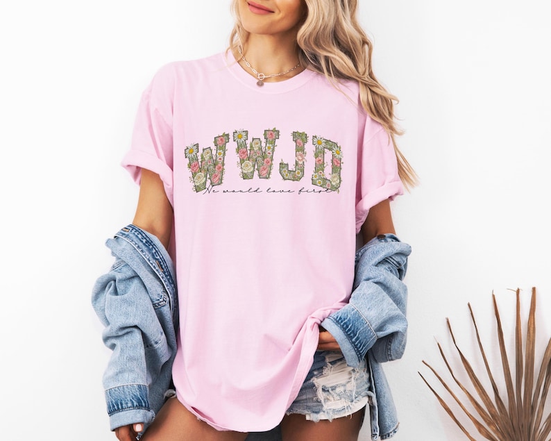 K&ouml;nnte beinhalten: Hellrosa T-Shirt mit den Buchstaben "WWJD" in floralem Design. Darunter steht der Text "No world love first". Das Shirt wird mit einer Jeansjacke und Jeansshorts getragen.