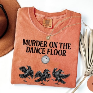 May include: Coral-colored t-shirt with the phrase "MURDER ON THE DANCE FLOOR" in black, featuring a disco ball graphic and three crow illustrations. A gold necklace and a brown hat are also visible.