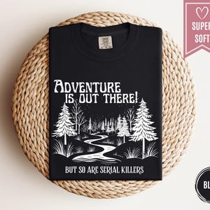 May include: Black t-shirt with white graphic text and illustration. The text reads "Adventure is out there! But so are serial killers." The illustration depicts a forest scene with trees and a winding river. The shirt is labeled "Super Soft."