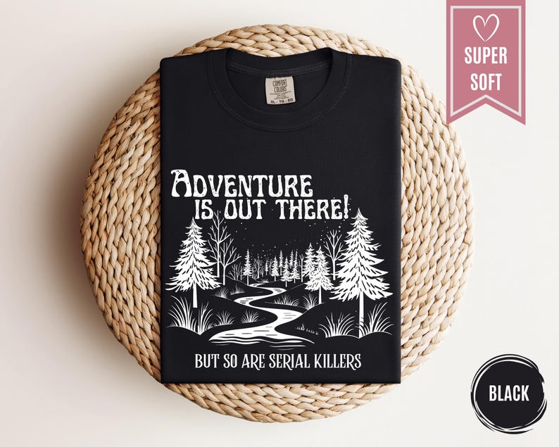 May include: Black t-shirt with white graphic text and illustration. The text reads "Adventure is out there! But so are serial killers." The illustration depicts a forest scene with trees and a winding river. The shirt is labeled "Super Soft."