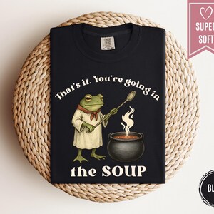 May include: Black t-shirt featuring a cartoon frog chef stirring a pot of soup. The text on the shirt reads "That's it. You're going in the SOUP." A pink banner with a heart and the words "SUPER SOFT" is in the upper right corner.